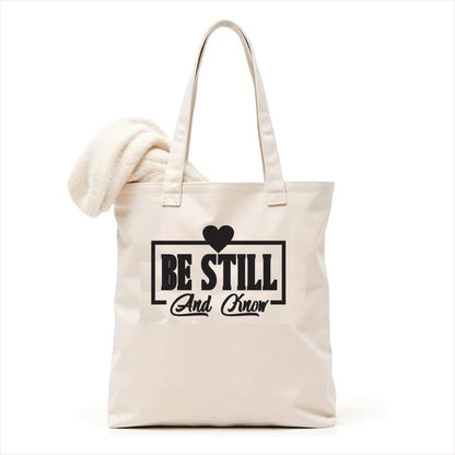 Beige tote bag with 'Be Still and Know' text on a white background