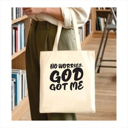 Person holding a tote bag with 'No worries, God got me' text in a library setting