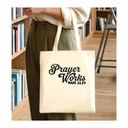 Tote bag with 'Prayer Works' text held by a person in a library setting