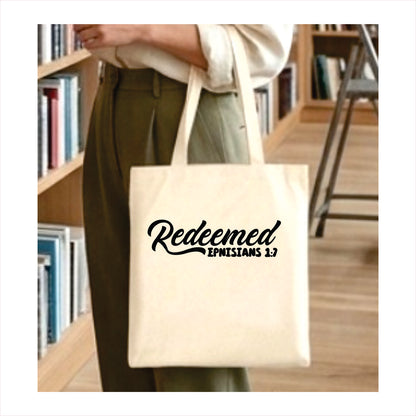 Person holding a tote bag with 'Redeemed Ephesians 1:7' text in a library setting.