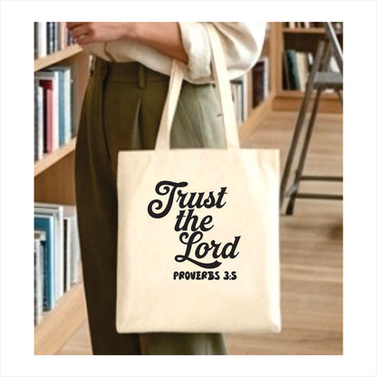 Tote bag with 'Trust the Lord Proverbs 3:5' text held by a person in a library setting.